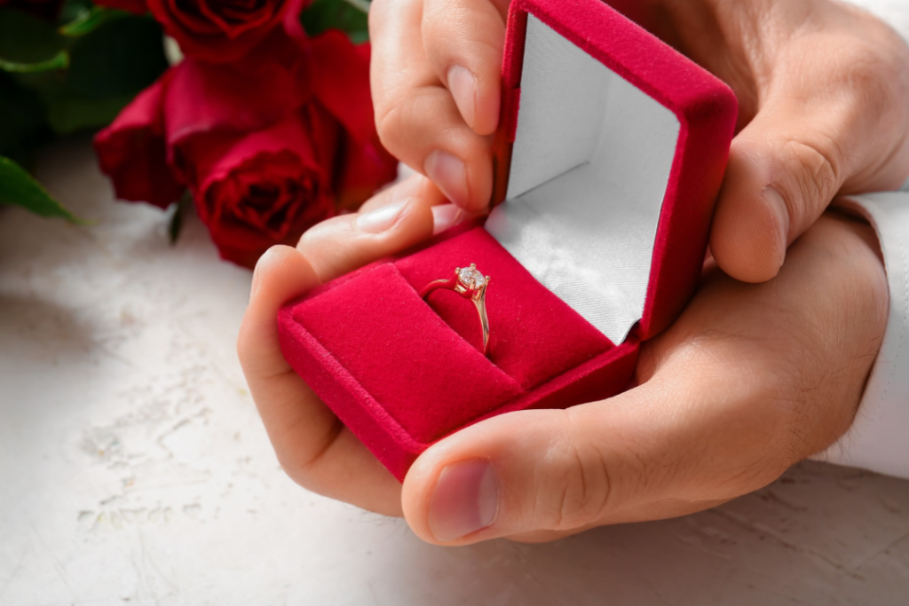 Mani di una persona che aprono una scatolina di velluto rosso con un anello solitario in oro e diamante, affiancata da rose rosse, per un regalo dal significato speciale.